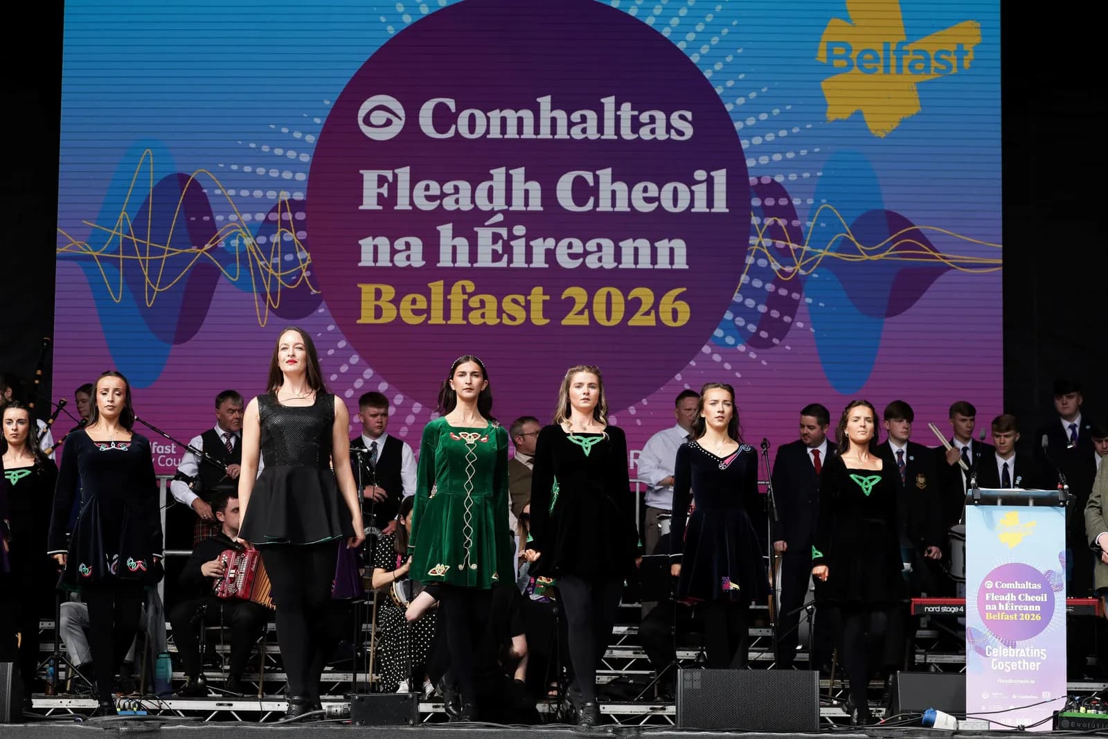 Several women in traditional Irish dance costumes stand on a stage in front of a large screen displaying the 'Comhaltas Fleadh Cheoil na hΓireann Belfast 2026' logo, with musicians and other performers in the background.
